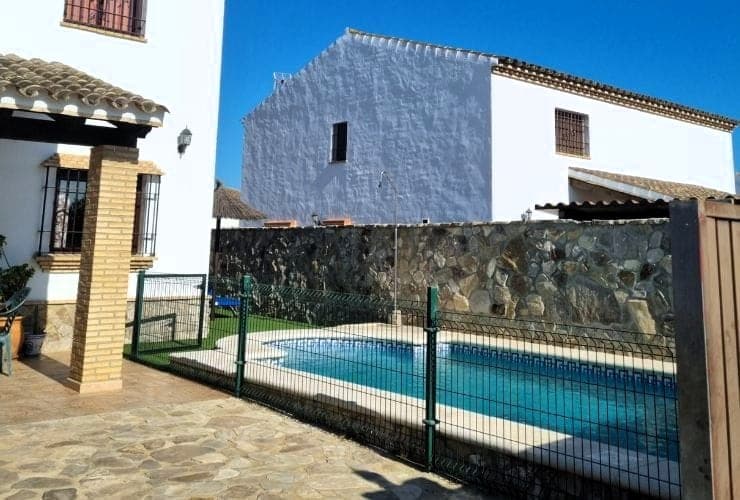 piscina vallada con verja verde, rodeada de suelo de piedra y muro de mampostería, junto a fachadas blancas con tejado de teja