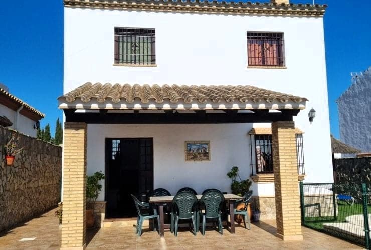 casa blanca de dos plantas con porche de columnas de ladrillo, mesa de madera y sillas verdes de plástico bajo cubierta de teja