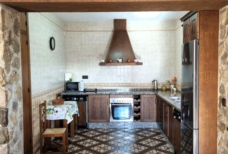 cocina en L con muebles de madera oscura, campana de madera sobre la placa, nevera de acero inoxidable y suelo de baldosas hidráulicas con mesa y sillas de paja al fondo