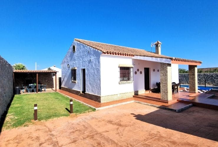 casa blanca de una planta con tejado de teja árabe, porche con columnas de ladrillo y patio exterior con zona de césped y pérgola con mesa y sillas a la izquierda