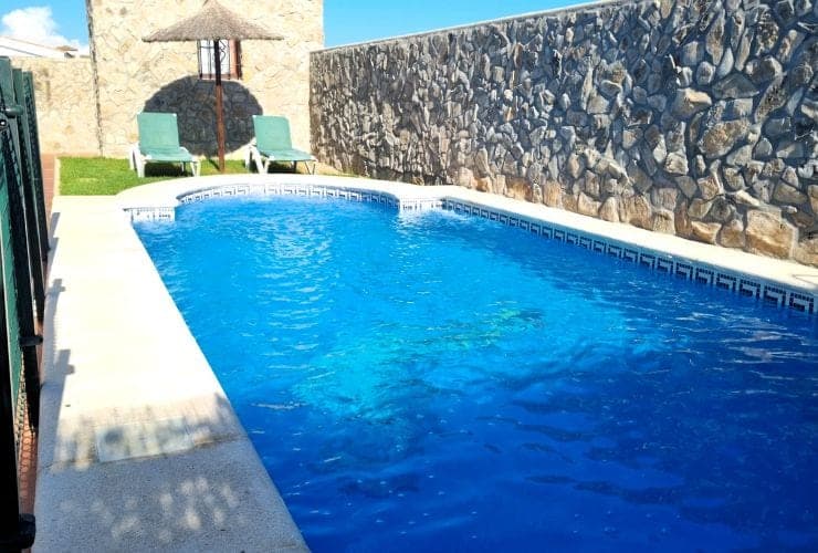 piscina rectangular con borde de baldosas decorativas, dos tumbonas verdes y sombrilla de paja junto a un muro de piedra
