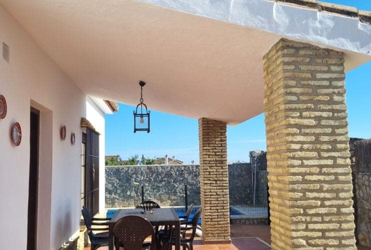 terraza cubierta con columnas de ladrillo, mesa de comedor con sillas y farol colgado, al fondo piscina tras un muro de piedra