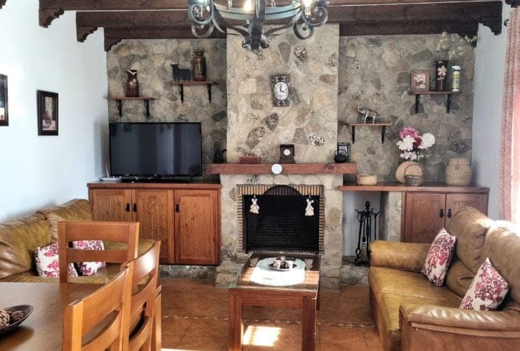 salón con chimenea de piedra, sofás de cuero marrón con cojines floridos, televisión sobre mueble de madera y mesa de comedor en primer plano
